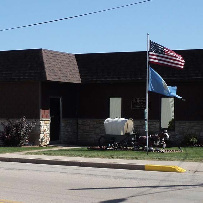 A small brown brick building with two flags—an American flag and a blue state flag—flying on a pole outside. Near the entrance, a miniature covered wagon display with mannequins adds charm to this spot near Sherwood Bar, known for some of the best food around.