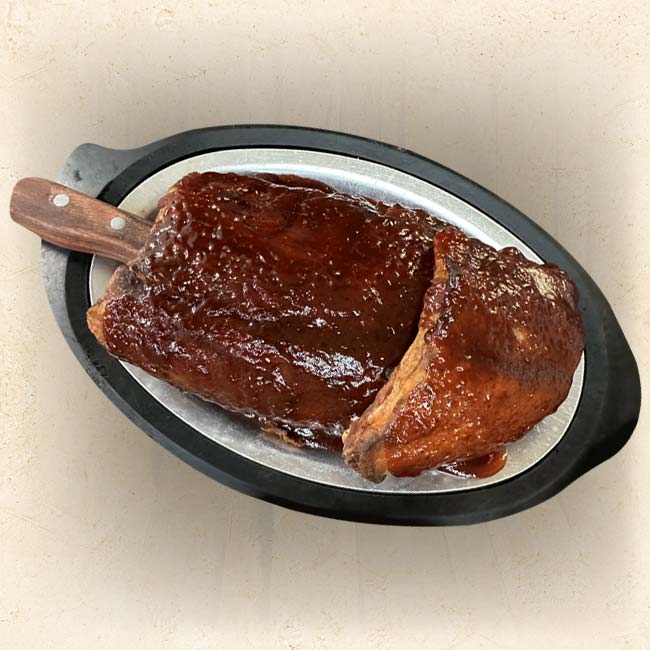A cooked brisket glazed with barbecue sauce is served on an oval metal platter with a handle, set inside a black serving tray at The Granary Supper Club—perfect for anyone searching for the Best Food in town. A knife is partially visible under the brisket.