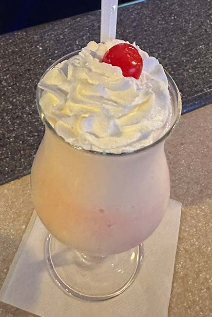 A creamy milkshake topped with whipped cream and a cherry, served in a curved glass with a straw at Sherwood Restaurant, placed on a white napkin on the countertop.