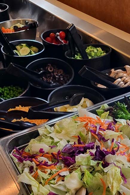 A vibrant salad bar at Sherwood Restaurant features trays of leafy greens, shredded carrots, and cabbage in the front, with bowls of the Best Food toppings like tomatoes, broccoli, olives, mushrooms, peas, and shredded cheese in the background.