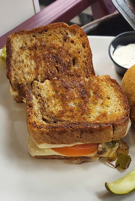 A close-up of a grilled sandwich halved on a white plate at Sherwood Restaurant, filled with tomato, cheese, and vegetables. A pickle spear and a small container of dipping sauce are visible in the background—enjoy the best food around!.