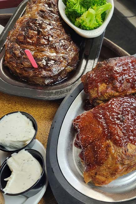 Two plates of grilled meat, one with a large steak and a side of broccoli, the other with glazed ribs, served at Sherwood Restaurant. Three small cups of creamy white dipping sauce sit nearby, adding to the Best Food experience.