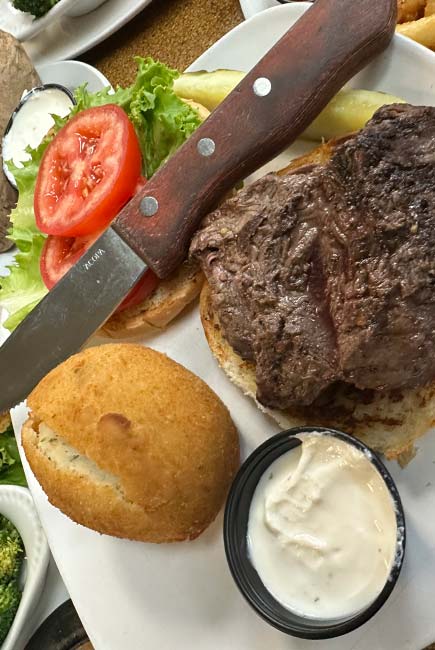 Enjoy the Best Food at Sherwood Bar—a steak sandwich with tomatoes, lettuce, a pickle spear, a biscuit, and white dipping sauce on the side. A knife rests atop the bun for easy sharing.