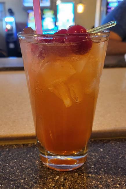 A tall glass filled with an orange iced cocktail, garnished with red cherries and a pink straw, sits on a speckled countertop at Sherwood Bar, inviting guests to unwind in its vibrant bar setting.