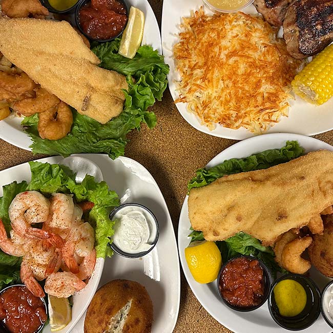 Four plates of seafood dishes at The Granary Supper Club, featuring fried fish, shrimp, a crab cake, tartar and cocktail sauces, lemon wedges, corn on the cob, and shredded hashbrowns—all served on a bed of lettuce for the Best Food experience.