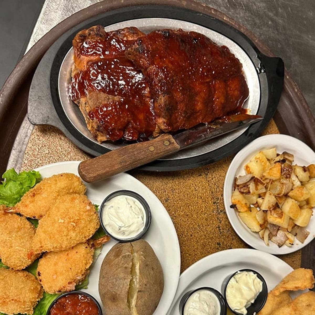 A platter of barbecued ribs with a knife, served with fried shrimp, dipping sauces, a baked potato, and a bowl of roasted potatoes on the side—enjoy this Best Food favorite at The Granary Supper Club in Sherwood Bar.