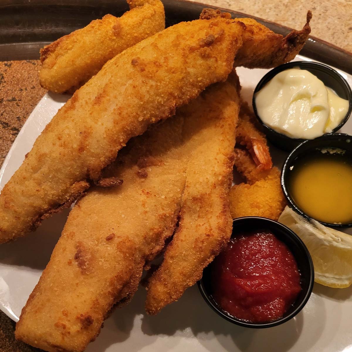 A plate of breaded and fried fish fillets with hush puppies, served with tartar sauce, melted butter, cocktail sauce, and lemon wedges—just like the Best Food at The Granary Supper Club.