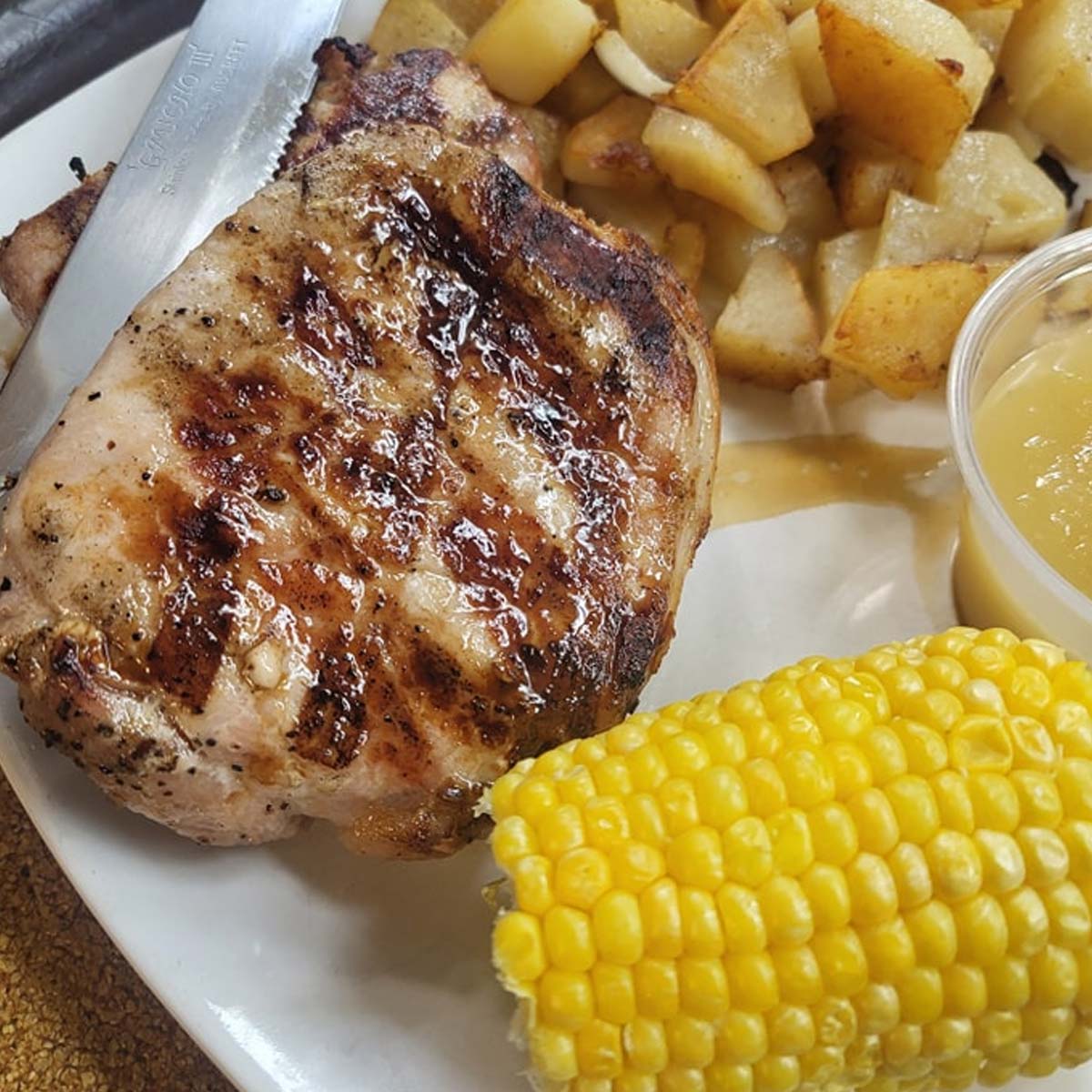 A plate at Sherwood Restaurant features a grilled pork chop, roasted diced potatoes, a piece of corn on the cob, and a small cup of applesauce. A knife rests on the pork chop, showcasing some of the Best Food around.