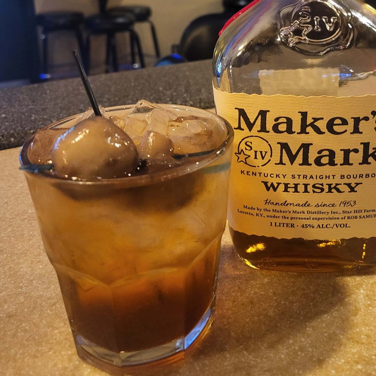 A glass filled with ice, dark liquid, and cocktail onions on a pick sits next to a bottle of Makers Mark Kentucky Straight Bourbon Whisky on the Sherwood Bar countertop.