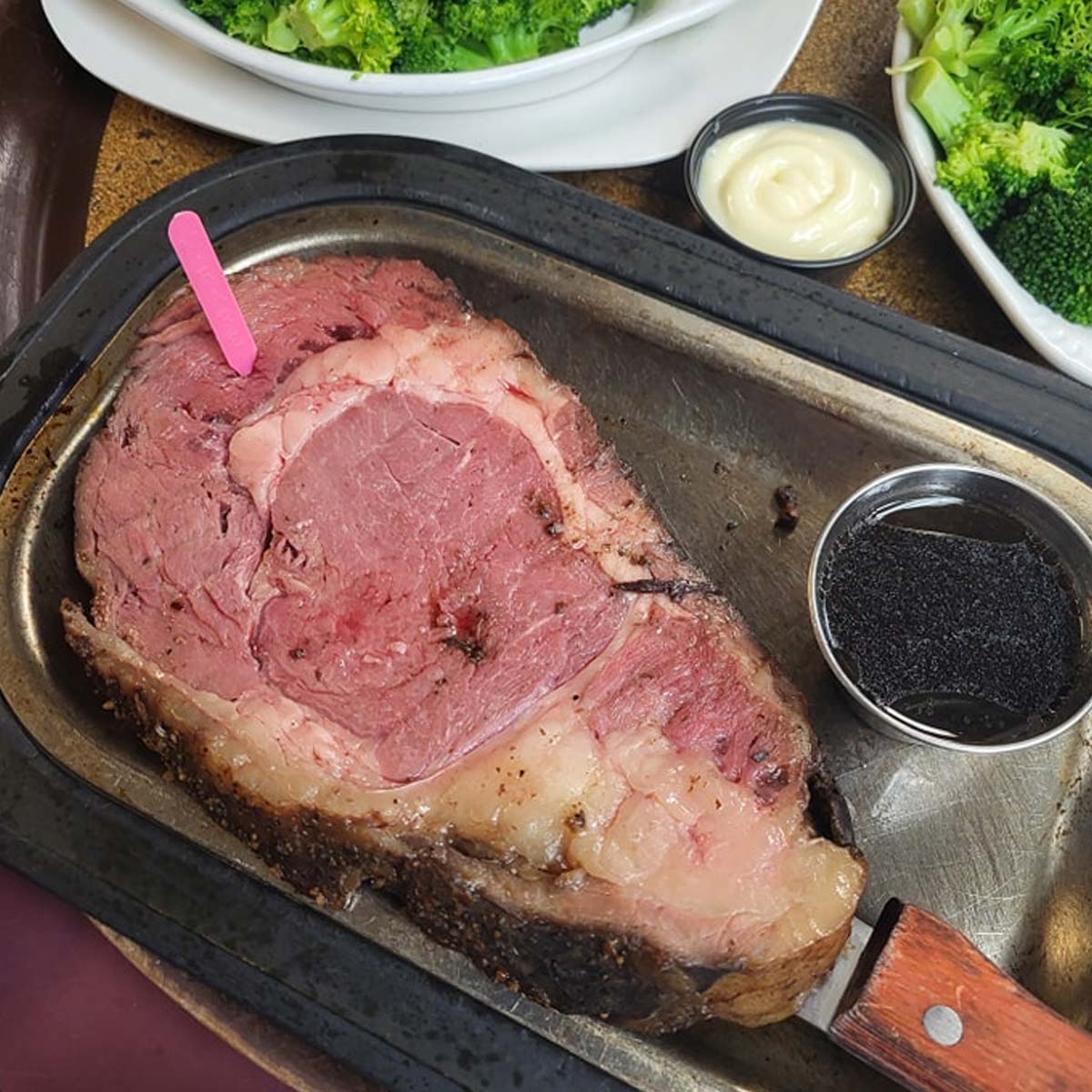 A large slice of rare prime rib on a metal tray, served with a knife, a cup of dark au jus, a pink doneness marker, and sides of broccoli and mayonnaise—just like youd find at The Granary Supper Club.