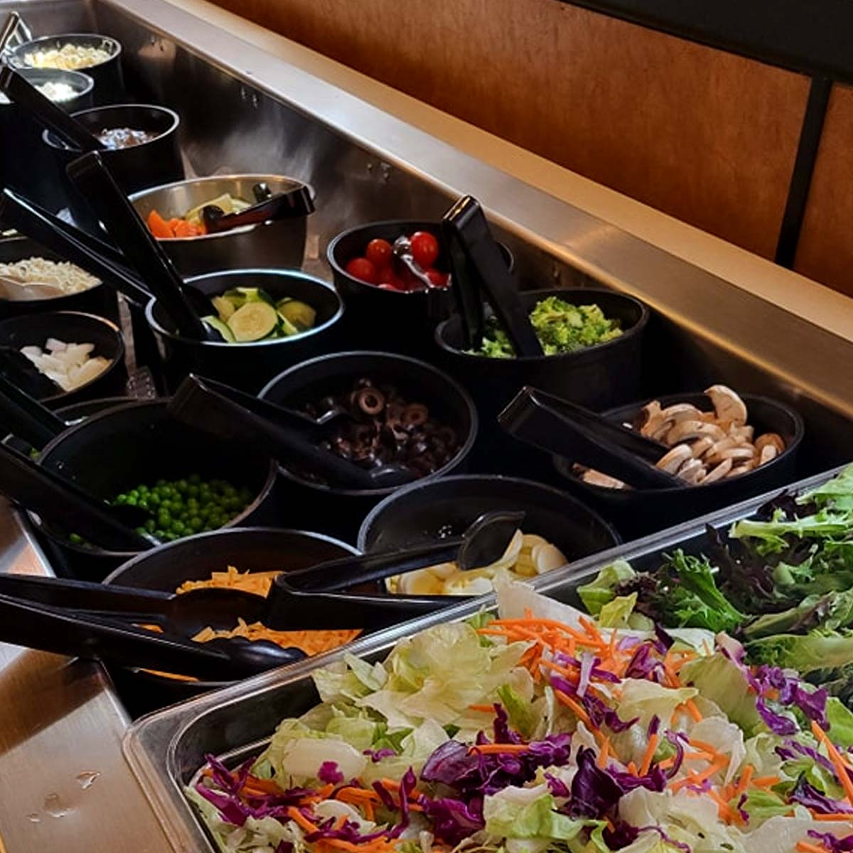 A salad bar at The Granary Supper Club features fresh ingredients like leafy greens, peas, mushrooms, and cheese in black bowls with serving tongs—perfect for those seeking the Best Food experience.