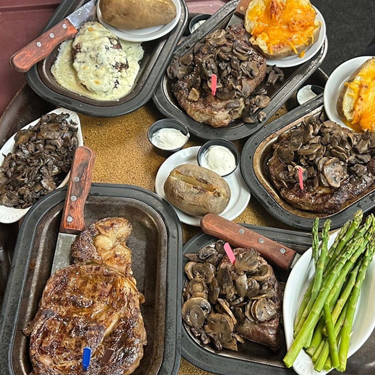 At Sherwood Restaurant, several plates of steak topped with sautéed mushrooms are arranged alongside baked potatoes, mashed potatoes with gravy, twice-baked potatoes, bowls of sour cream, and grilled asparagus for the best food experience.