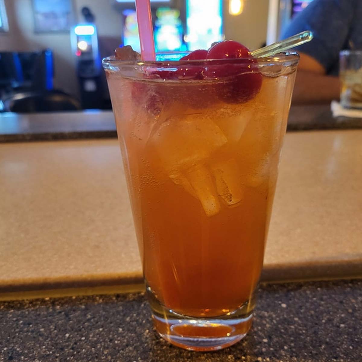A tall glass filled with an orange-brown iced drink, topped with three red cherries and served with a straw, sits on a countertop at Sherwood Bar, capturing the inviting ambiance of one of the best food spots in town.
