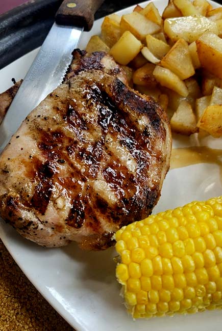 Grilled pork chop with char marks, served with a knife, golden brown diced potatoes, and a piece of corn on the cob on a white plate—a classic at The Granary Supper Club, known for the Best Food in town.