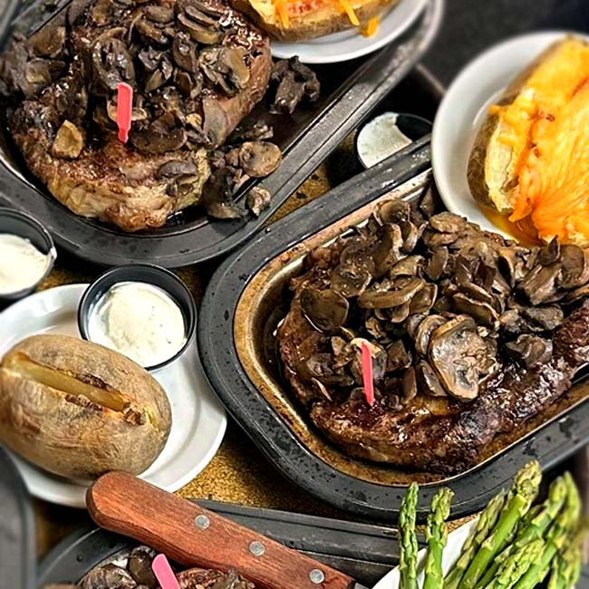 Two plates of steak topped with sautéed mushrooms, served with baked potatoes, sour cream, cheese, and asparagus are beautifully arranged on metal trays at The Granary Supper Club. A knife rests on the side for a classic dining experience.