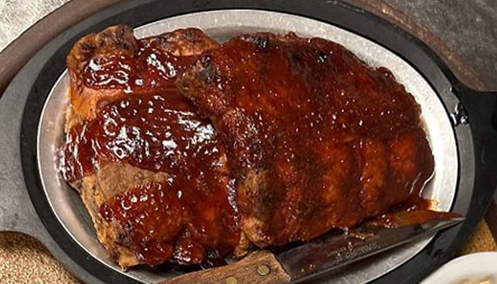 A platter of glazed barbecued ribs, coated in a rich, shiny barbecue sauce, served at The Granary Supper Club with a carving knife placed beside the ribs on the metal tray—truly some of the best food around.