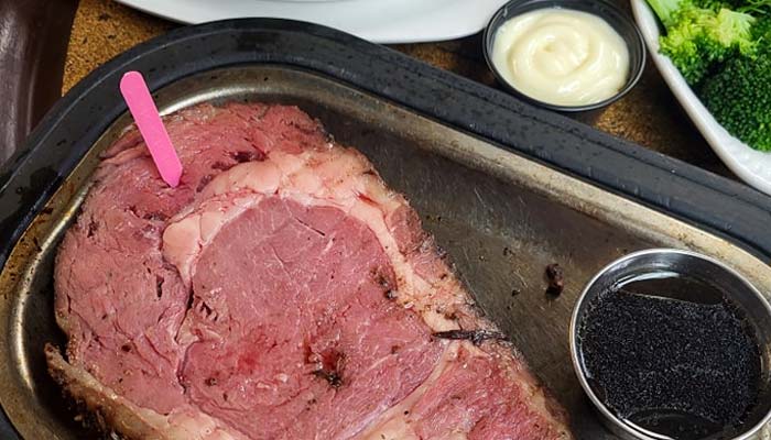 A thick slice of rare prime rib on a metal tray with a pink doneness marker, served with au jus, creamy horseradish sauce, and steamed broccoli—classic fare at Sherwood Restaurant for those seeking the best food in town.