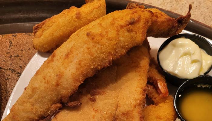 A plate of golden-brown fried fish fillets with hush puppies, served with creamy tartar sauce and melted butter—a classic from Sherwood Restaurant, known for offering the best food in town.