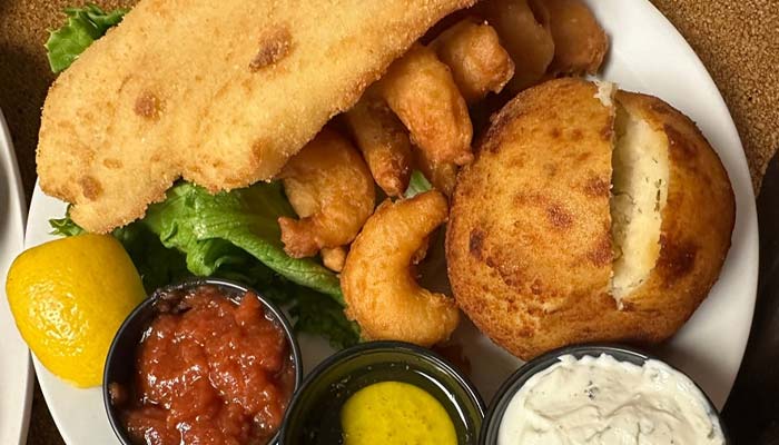 A plate of fried seafood from The Granary Supper Club features breaded fish, shrimp, a split hushpuppy, lettuce, a lemon wedge, and cups of cocktail sauce, tartar sauce, and another dipping sauce—experience the best food in town!.