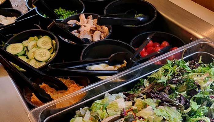 At Sherwood Bar, enjoy a vibrant salad bar with black bowls filled with sliced cucumbers, shredded cheese, cherry tomatoes, peas, mushrooms, and mixed greens—each with its own serving tongs. Experience the best food at Sherwood Restaurant.