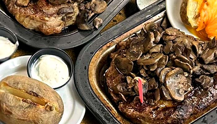 Grilled steaks topped with sautéed mushrooms on metal trays, served with baked potatoes and small containers of sour cream—experience the best food at The Granary Supper Club.