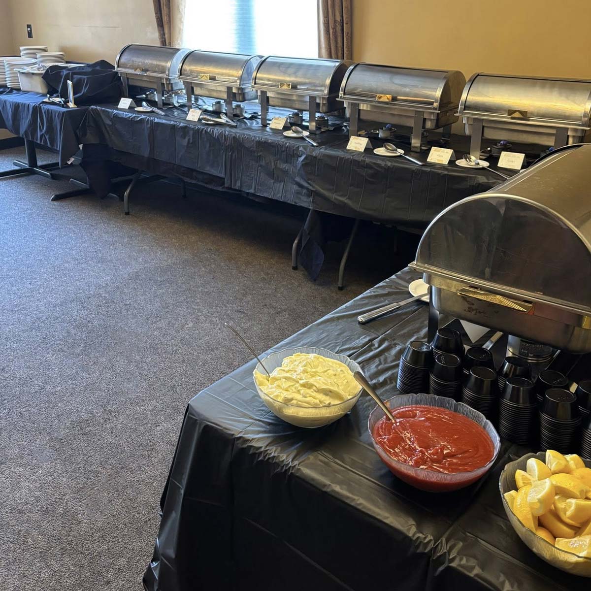 A buffet setup with covered chafing dishes lines tables draped in black cloth at The Granary Supper Club. In the foreground are bowls of whipped topping and red sauce, lemon wedges, plastic cups, and utensils.