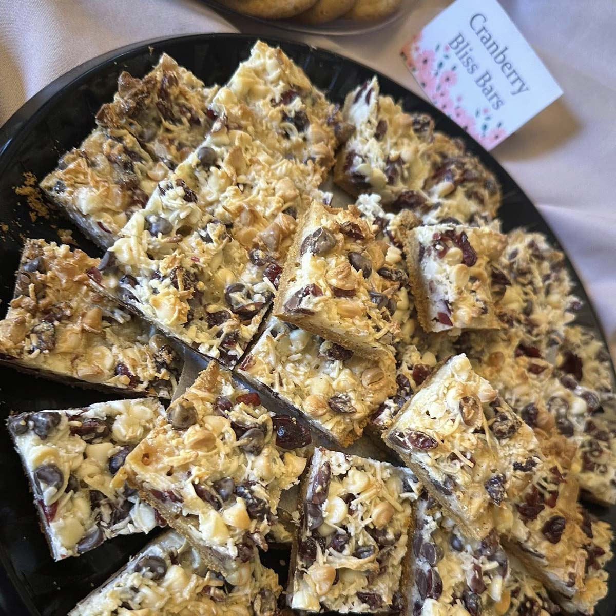 A plate filled with cranberry bliss bars cut into squares, topped with white chocolate chips, cranberries, and nuts. In the background, a small sign reads Cranberry Bliss Bars at Sherwood Restaurant.