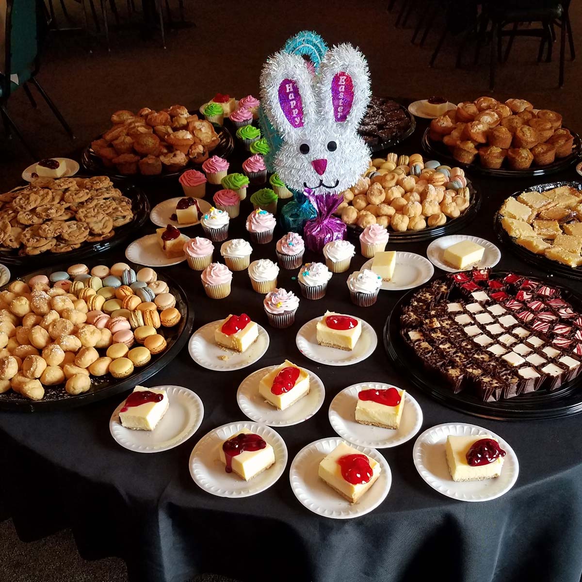 A table filled with assorted desserts, including cupcakes, cookies, pastries, and cheesecake slices topped with fruit from Sherwood Restaurant. A decorative bunny centerpiece with Happy Birthday signs is in the middle.