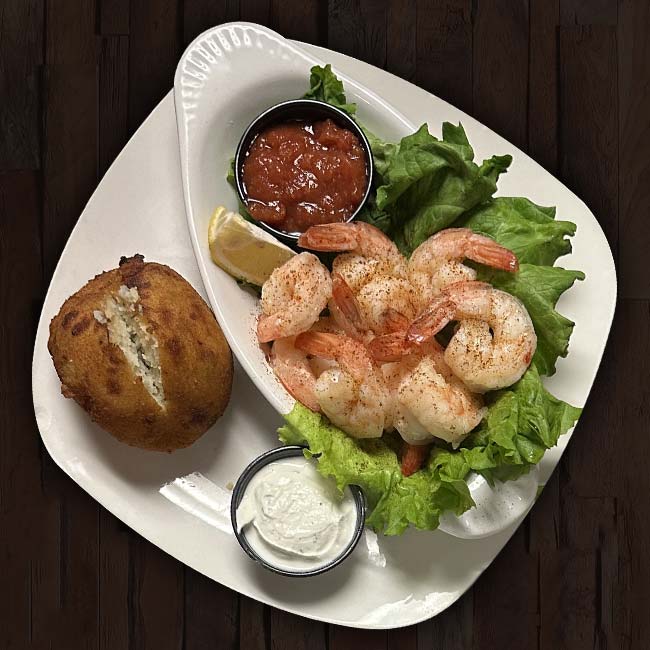 A white plate with shrimp on lettuce, a lemon wedge, cocktail sauce, a breaded stuffed crab cake, and creamy dipping sauce, set on a dark wooden table at Sherwood Bar—home of the best food in town.