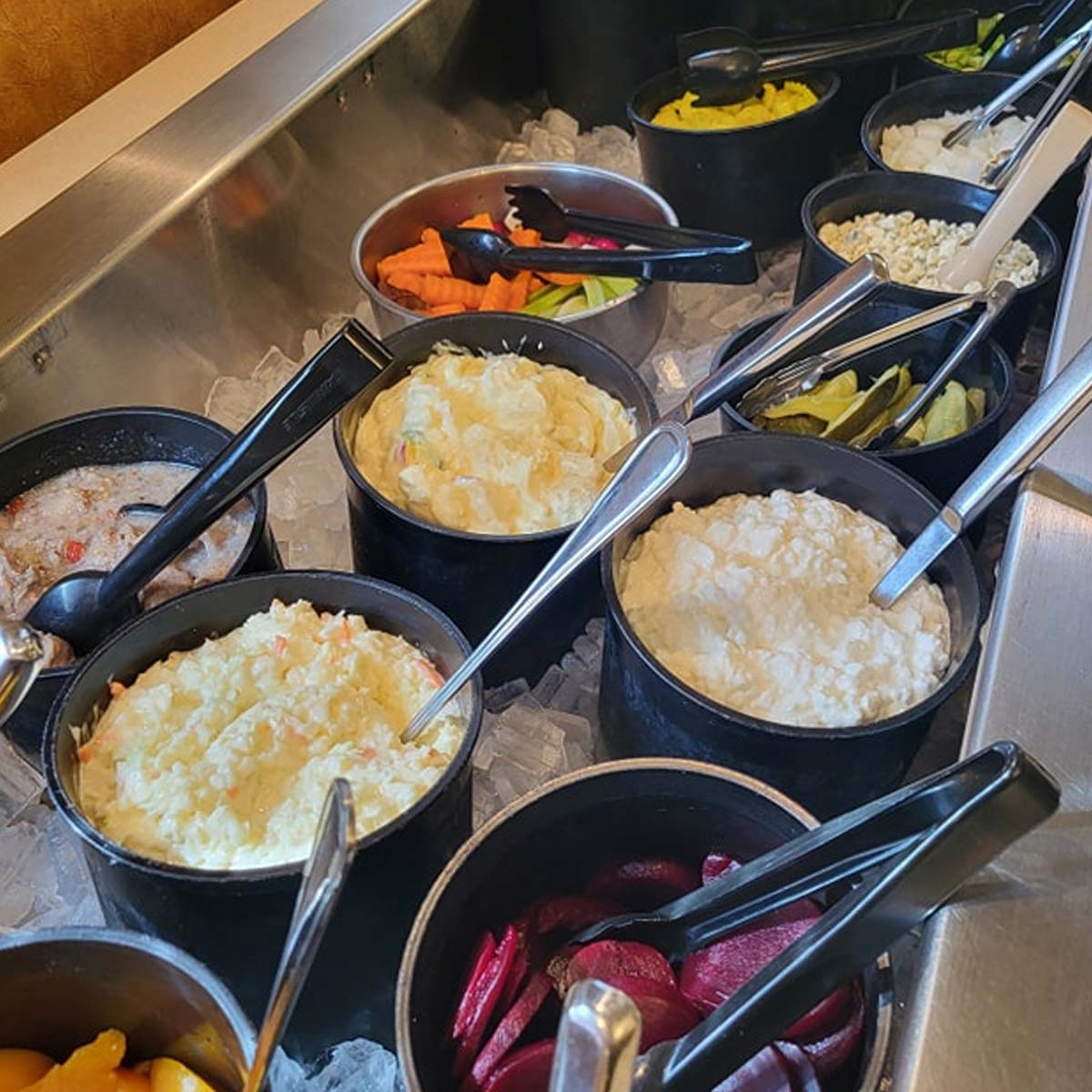 A salad bar at Sherwood Restaurant features black bowls filled with potato salad, pickled beets, cottage cheese, carrot sticks, lime wedges, and other fresh vegetables, all set on ice with serving utensils.