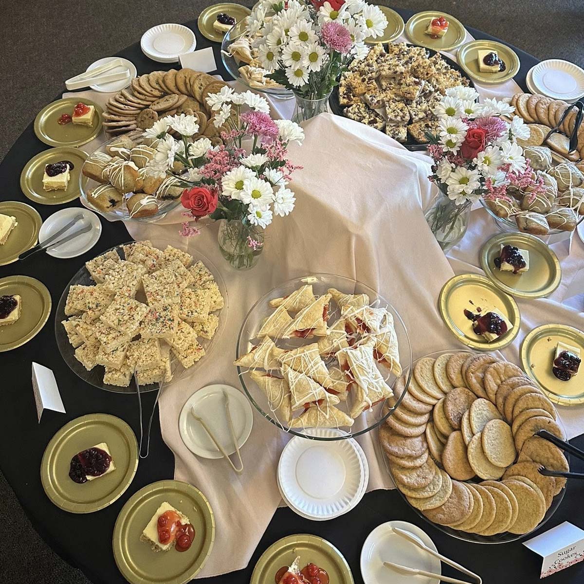 A round table set with assorted cookies, pastries, triangular sandwiches, and fruit-topped desserts, much like the elegant spreads at Sherwood Restaurant, features bouquets of white and pink flowers with plates and utensils arranged neatly.