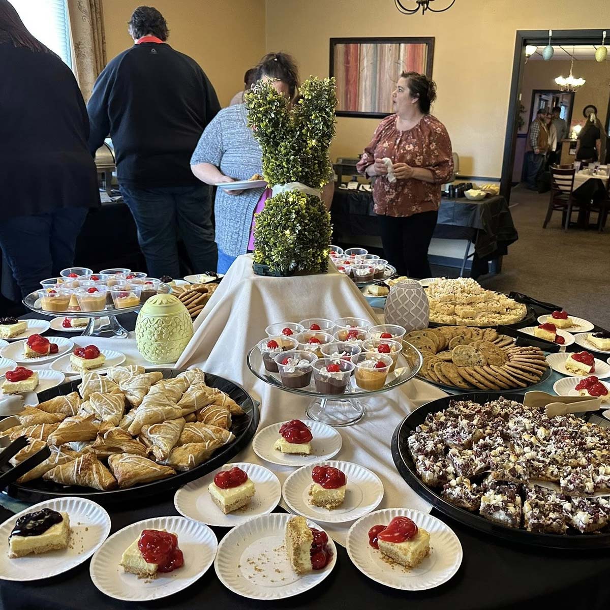 A large table at The Granary Supper Club is covered with various desserts, including cookies, pastries, cheesecakes with cherries, and parfait cups. Several people serve themselves in a warmly lit room adorned with framed art on the wall.
