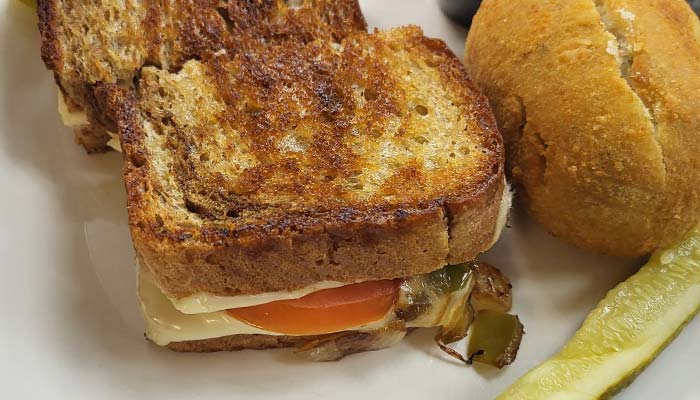 A toasted sandwich with melted cheese, tomato, and vegetables is on a plate next to a bread roll and a pickle spear—classic Best Food you’d expect from Sherwood Bar or The Granary Supper Club.