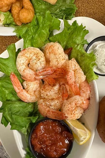 A plate of cooked shrimp on a bed of green leaf lettuce, sprinkled with seasoning, served with a lemon wedge, cocktail sauce, and a side of tartar sauce at The Granary Supper Club. Fried food is visible in the background.